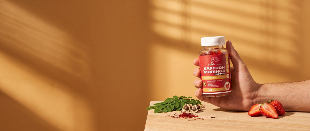 Hand holding Healthier Options Saffron and Moringa gummies in warm window light with moringa leaves and strawberries