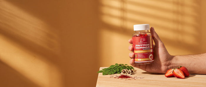 Hand holding Healthier Options Saffron and Moringa gummies in warm window light with moringa leaves and strawberries